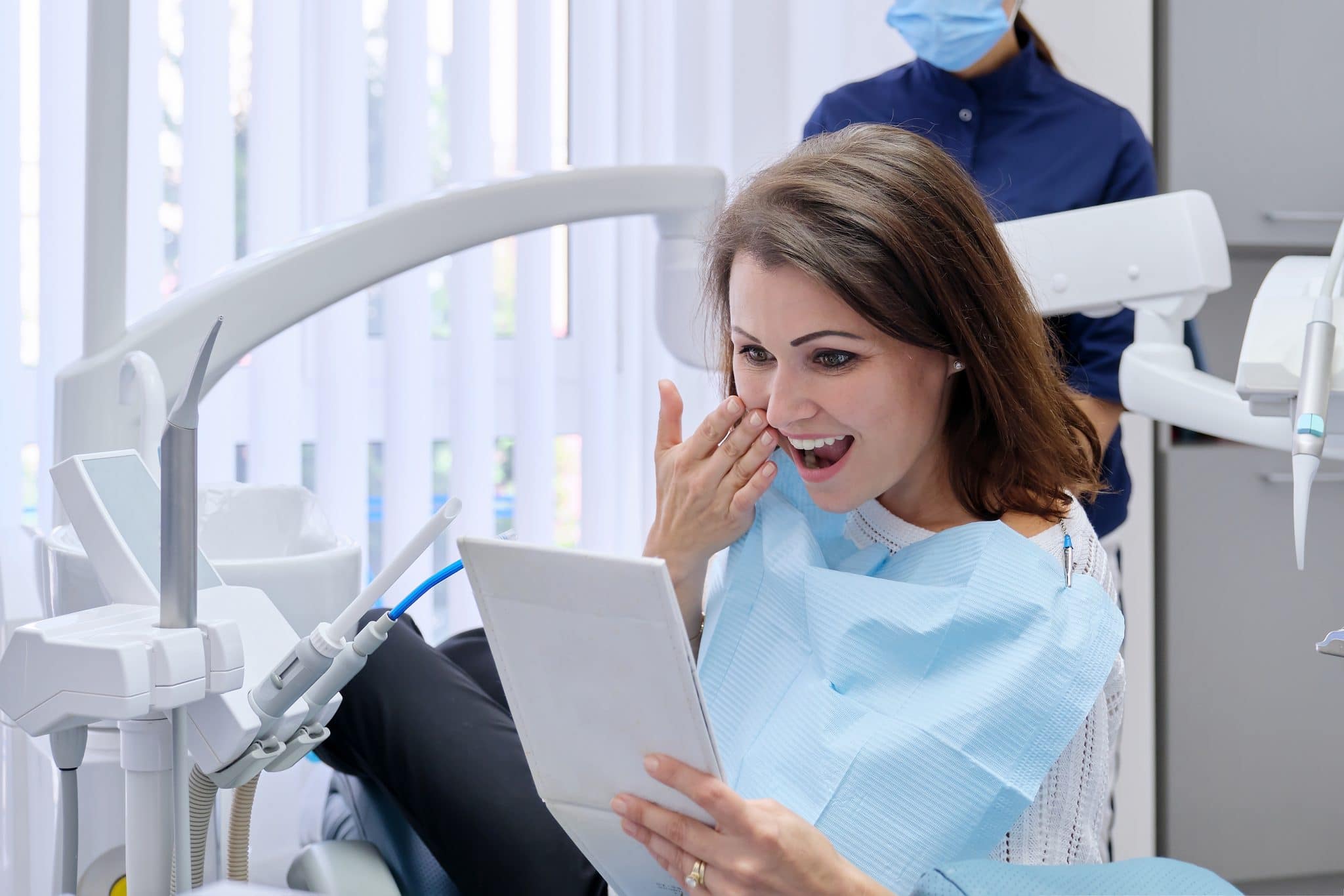 Happy middle aged woman with doctor dentist looking in mirror at teeth, sitting in dental chair. Medicine, dentistry and health care concept